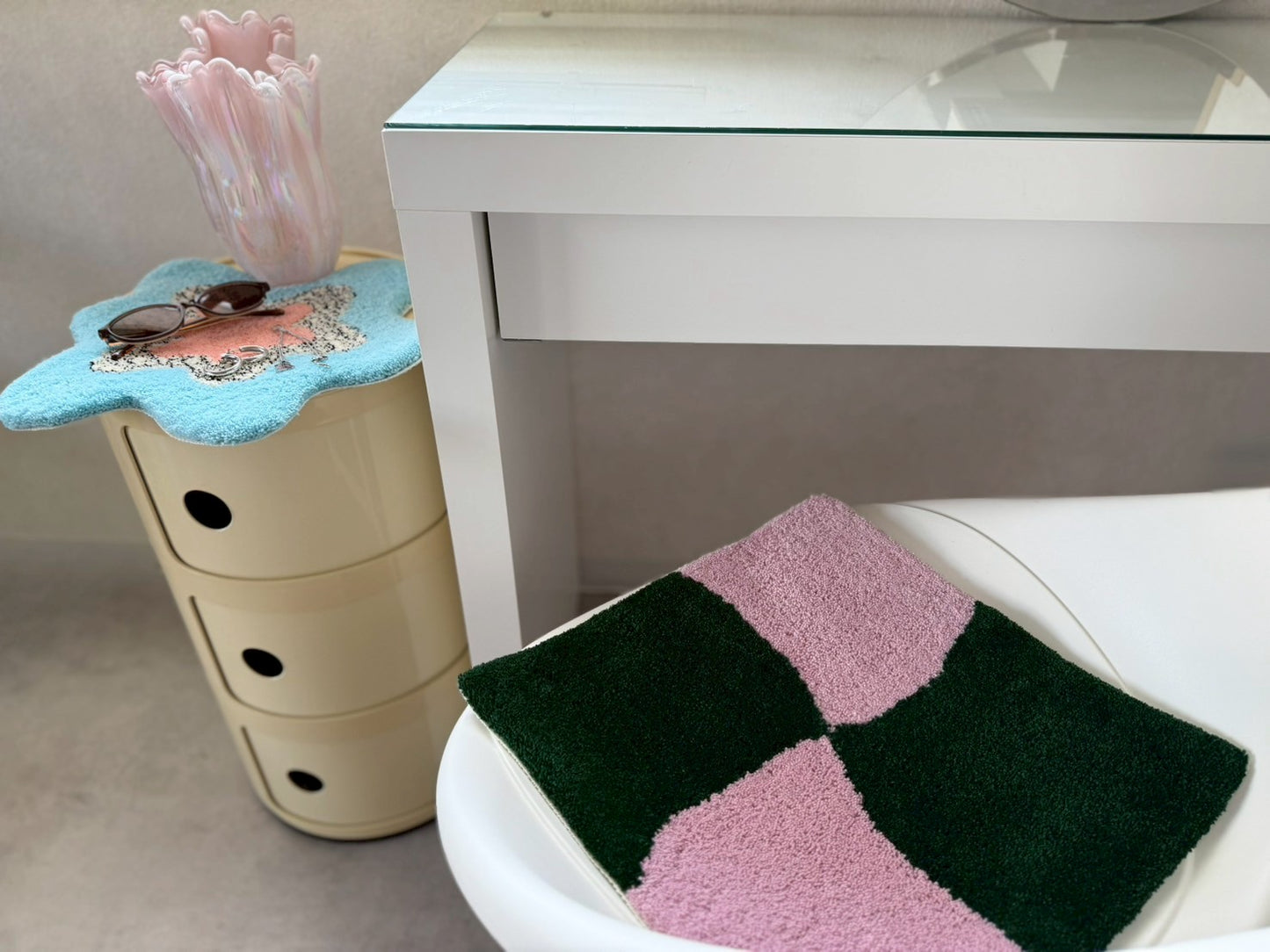 白い椅子とガラス天板のデスクのそばに置かれた2枚のタフティングラグ。椅子にはピンクと深緑の四角いデザイン、丸い収納棚の上には青とピンクの花型のラグが置かれている。チェア&アートラグコース タフティングワークショップ 大阪北堀江