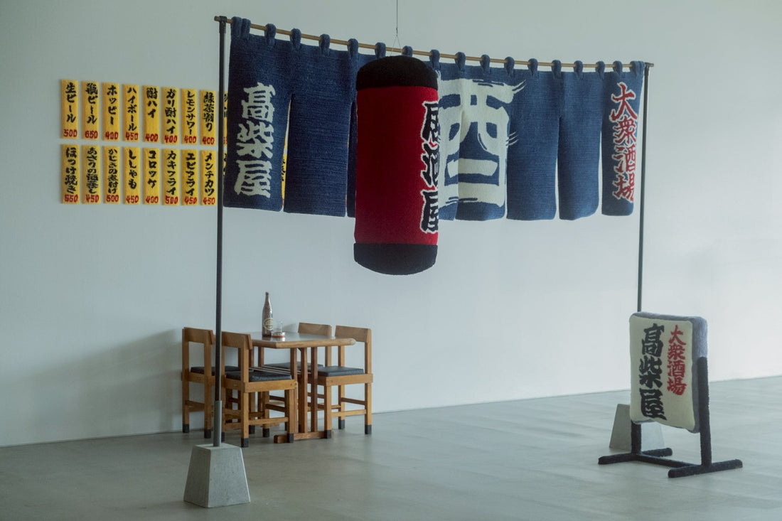 「居酒屋」をテーマにしたタフティングアート作品。暖簾（のれん）、提灯、看板、壁のメニュー表などがすべて毛糸で作られており、リアルな居酒屋の雰囲気を再現している。赤い提灯には「居酒屋」、のれんには「高楽屋」や「酒」の文字がタフティングで表現されている。奥には実際の木製テーブルと椅子が配置され、空間全体にリアリティとユーモアが融合したインスタレーション作品となっている。