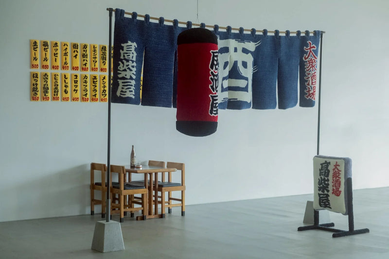 「居酒屋」をテーマにしたタフティングアート作品。暖簾（のれん）、提灯、看板、壁のメニュー表などがすべて毛糸で作られており、リアルな居酒屋の雰囲気を再現している。赤い提灯には「居酒屋」、のれんには「高楽屋」や「酒」の文字がタフティングで表現されている。奥には実際の木製テーブルと椅子が配置され、空間全体にリアリティとユーモアが融合したインスタレーション作品となっている。