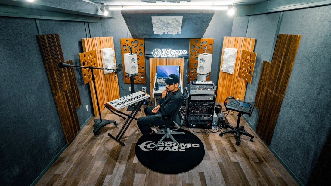 この画像は、音楽スタジオの内部を示しているようです。壁には音響パネルが設置され、中央にはモニターやキーボード、その他の音響機材が並べられています。