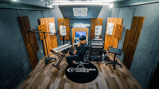 この画像は、音楽スタジオの内部を示しているようです。壁には音響パネルが設置され、中央にはモニターやキーボード、その他の音響機材が並べられています。