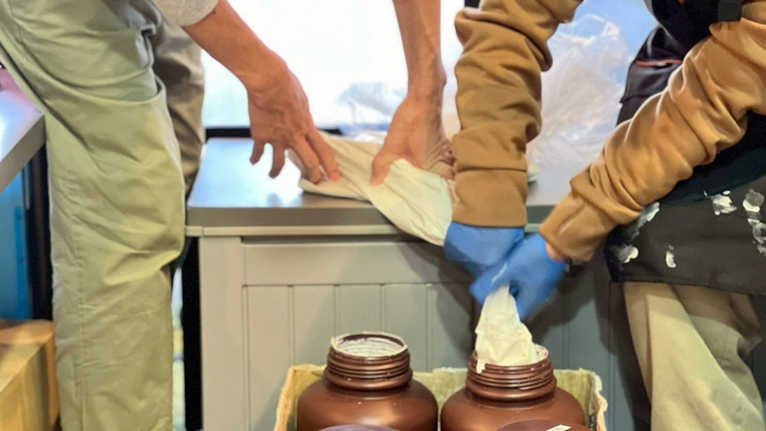 2人のスタッフが協力して白い素材の袋を持ち、茶色のボトルへ詰め替えている様子。手前には複数のボトルが並び、共同作業の温かい雰囲気が伝わる