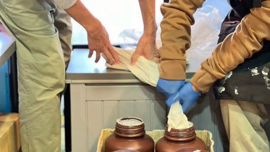 2人のスタッフが協力して白い素材の袋を持ち、茶色のボトルへ詰め替えている様子。手前には複数のボトルが並び、共同作業の温かい雰囲気が伝わる