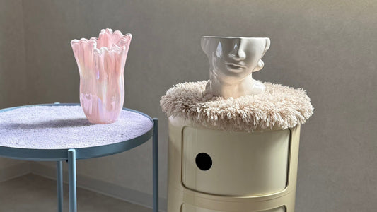 Kartellのコンポニビリの上にタフティングで作られたベージュの円形マットを敷き、上に白い彫刻のような顔型の花器を置いた様子。左側にはIKEAのグラドムの天板にラベンダーカラーのタフティングマットが敷かれ、上にピンクのガラス製フラワーベースが飾られている。モダンで洗練されたインテリア空間。