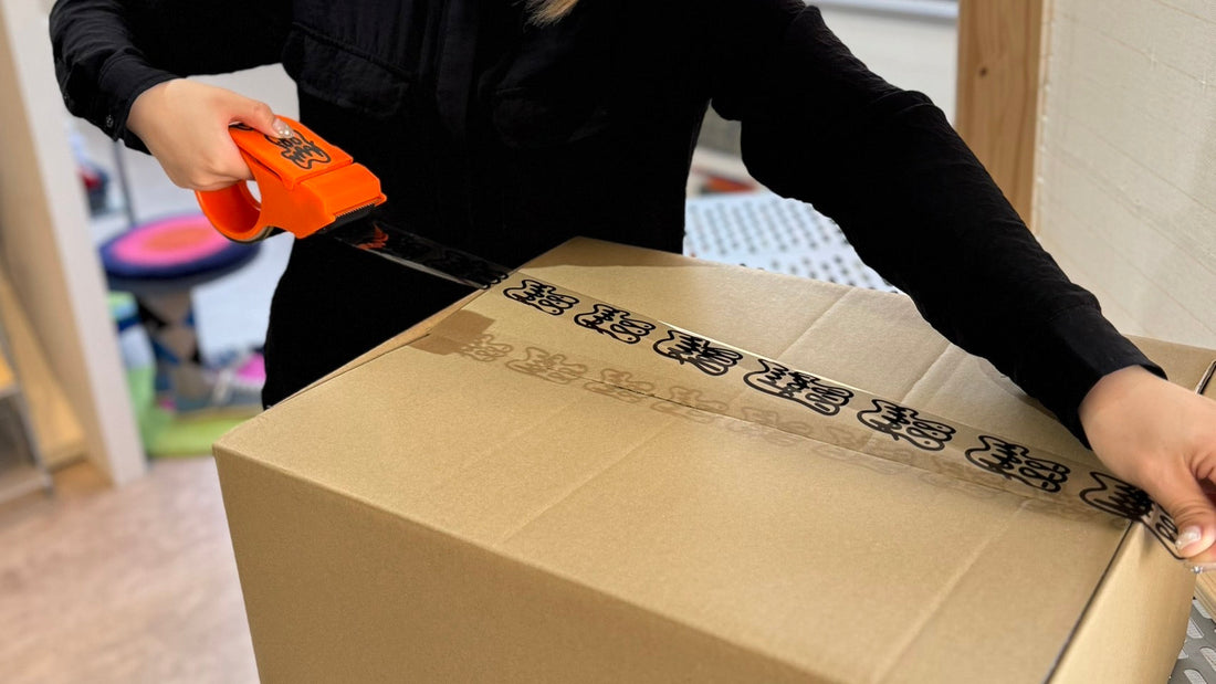 オレンジ色のテープディスペンサーでロゴ入りテープを貼り、段ボール箱を封している人物の手元のクローズアップ。