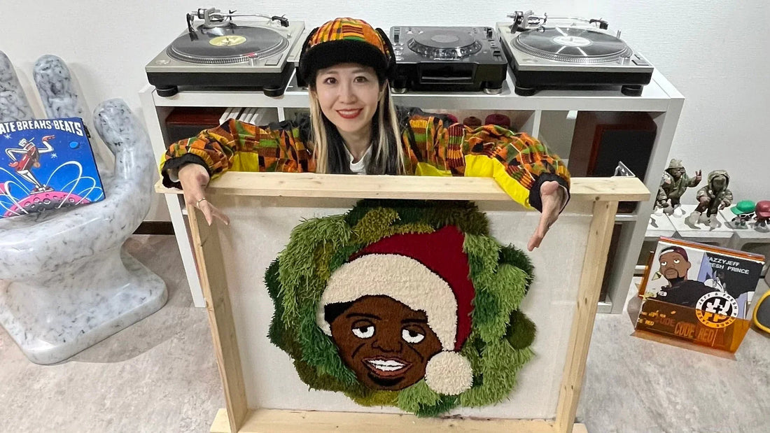 鮮やかな衣装を着たRISAさんが、サンタ帽をかぶったキャラクターが描かれたタフティング作品を持って笑顔でポーズをとっている。背景にはターンテーブルやレコードなど音楽関連のアイテムが並んでいる。