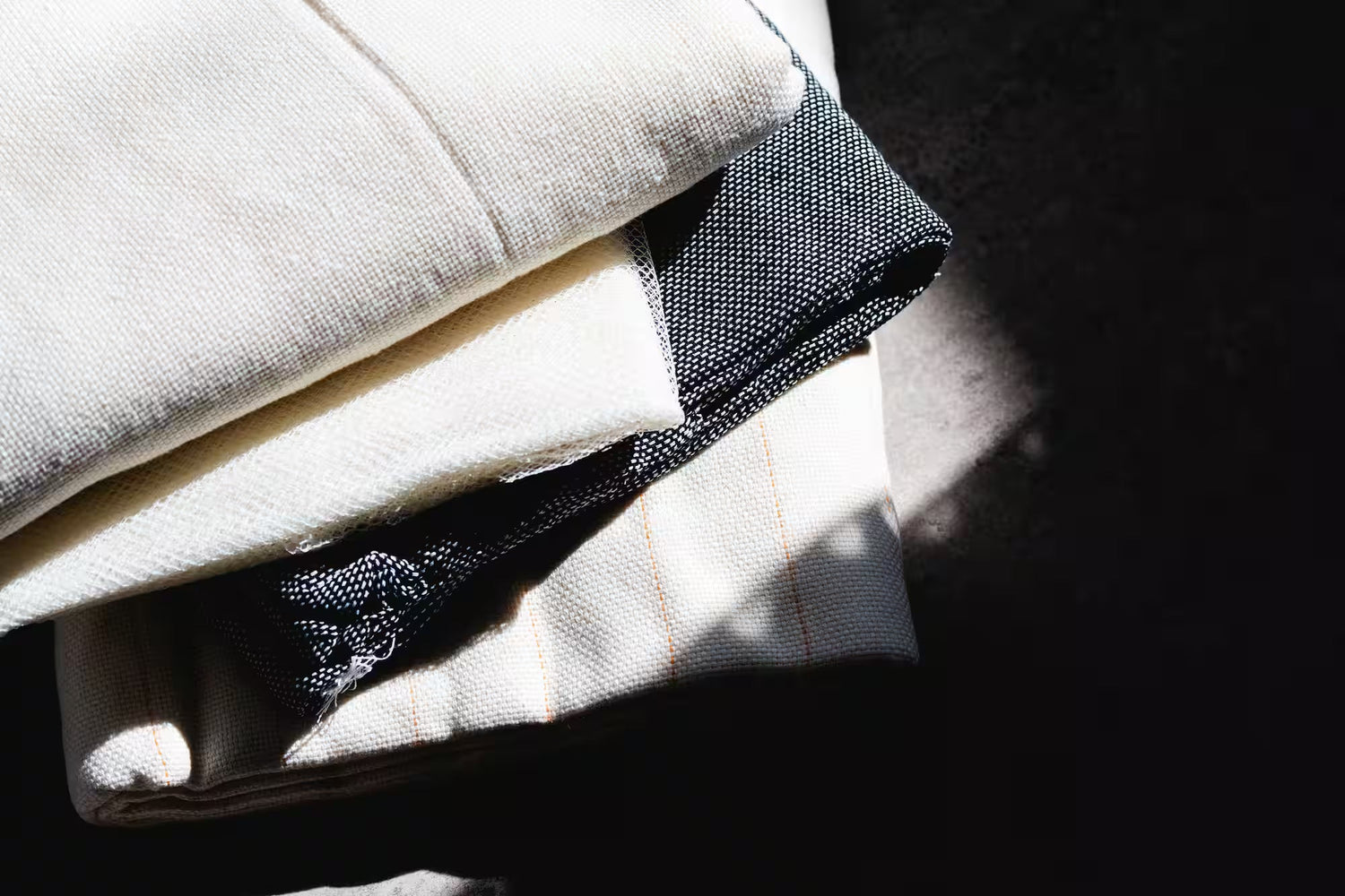  日差しの中で重ねられた布の束。白と黒の織り目の異なる布地が積み重ねられ、シンプルで質感の良さが感じられるデザイン。