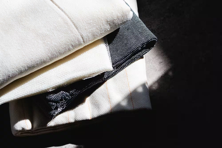  日差しの中で重ねられた布の束。白と黒の織り目の異なる布地が積み重ねられ、シンプルで質感の良さが感じられるデザイン。