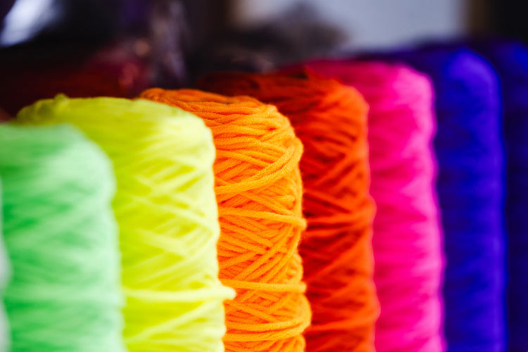 カラフルな毛糸が整然と並べられた棚。青、緑、赤、ピンク、オレンジ、黄色、茶色など、様々な色の毛糸がディスプレイされており、豊富なバリエーションが特徴的。