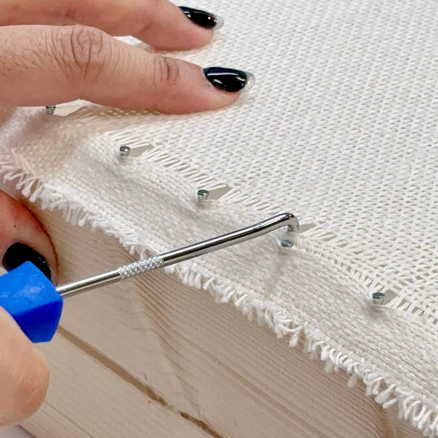 タフティング用の布をフレームに張る際に使用するフックツール（ツールピック）の使用例。
青い持ち手のツールで布目を引っかけ、ピンにきれいに固定している様子。布を均一に張ることで、タフティングガンでの打ち込みがスムーズに行える。