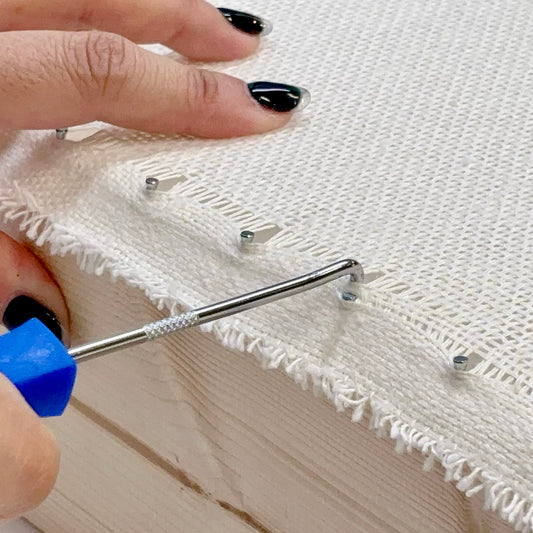 タフティング用の布をフレームに張る際に使用するフックツール（ツールピック）の使用例。
青い持ち手のツールで布目を引っかけ、ピンにきれいに固定している様子。布を均一に張ることで、タフティングガンでの打ち込みがスムーズに行える。
