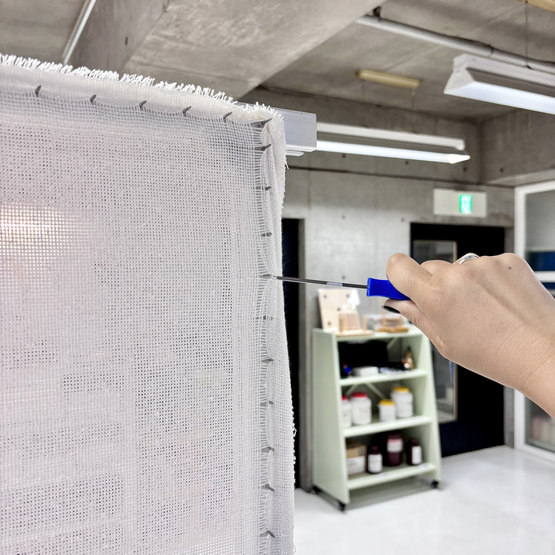 アルミフレームにタフティング用の基布を張り、青い持ち手のツールで端を押さえて調整している様子。背景には棚や作業道具が置かれた店頭の一角が映っている。