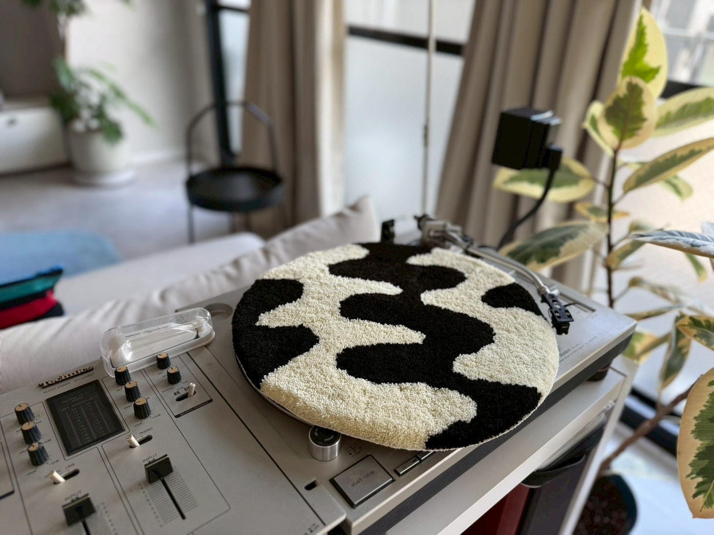 黒と白の模様が描かれた円形のタフティングラグ。のターンテーブルの上に置かれている。背景には観葉植物と部屋のインテリアが見える。チェア&アートラグコース タフティングワークショップ 大阪北堀江