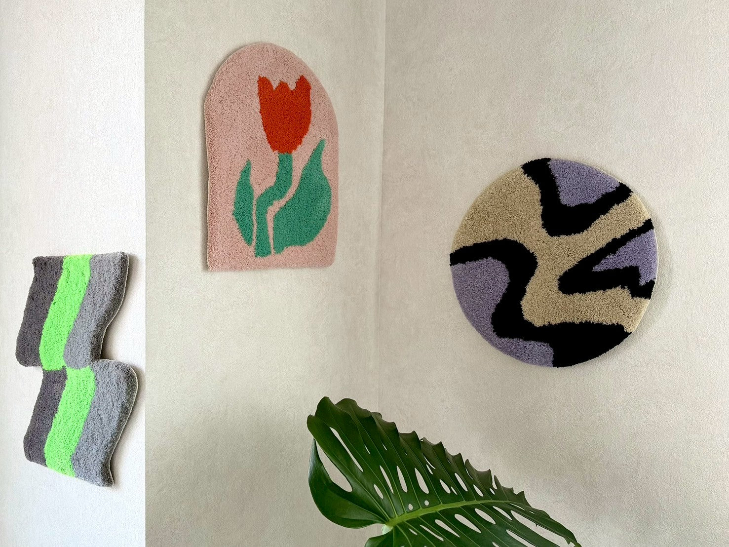 壁に飾られた3枚のタフティングラグ。赤いチューリップの絵柄があるアーチ型、紫とベージュに黒の模様をあしらった円形、そして灰色と緑のストライプ模様のラグ。チェア&アートラグコース タフティングワークショップ 大阪北堀江