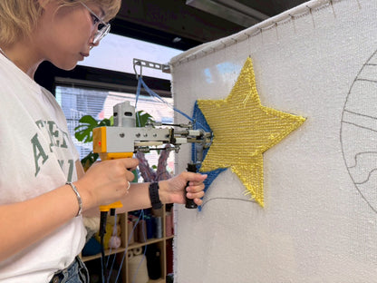 タフティングガンを持った人物が白い基布に黄色の星のラグを打ち込んでいる様子。背景には青い毛糸が見える。チェア&アートラグコース タフティングワークショップ 大阪北堀江