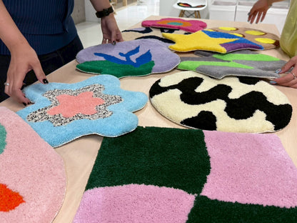 テーブルの上に並べられた、さまざまな形や模様のカラフルなタフティングラグ。花や星、幾何学模様などのデザインが見える。複数の人物が手でラグを触りながら制作するデザインを決めている。チェア&アートラグコース タフティングワークショップ 大阪北堀江